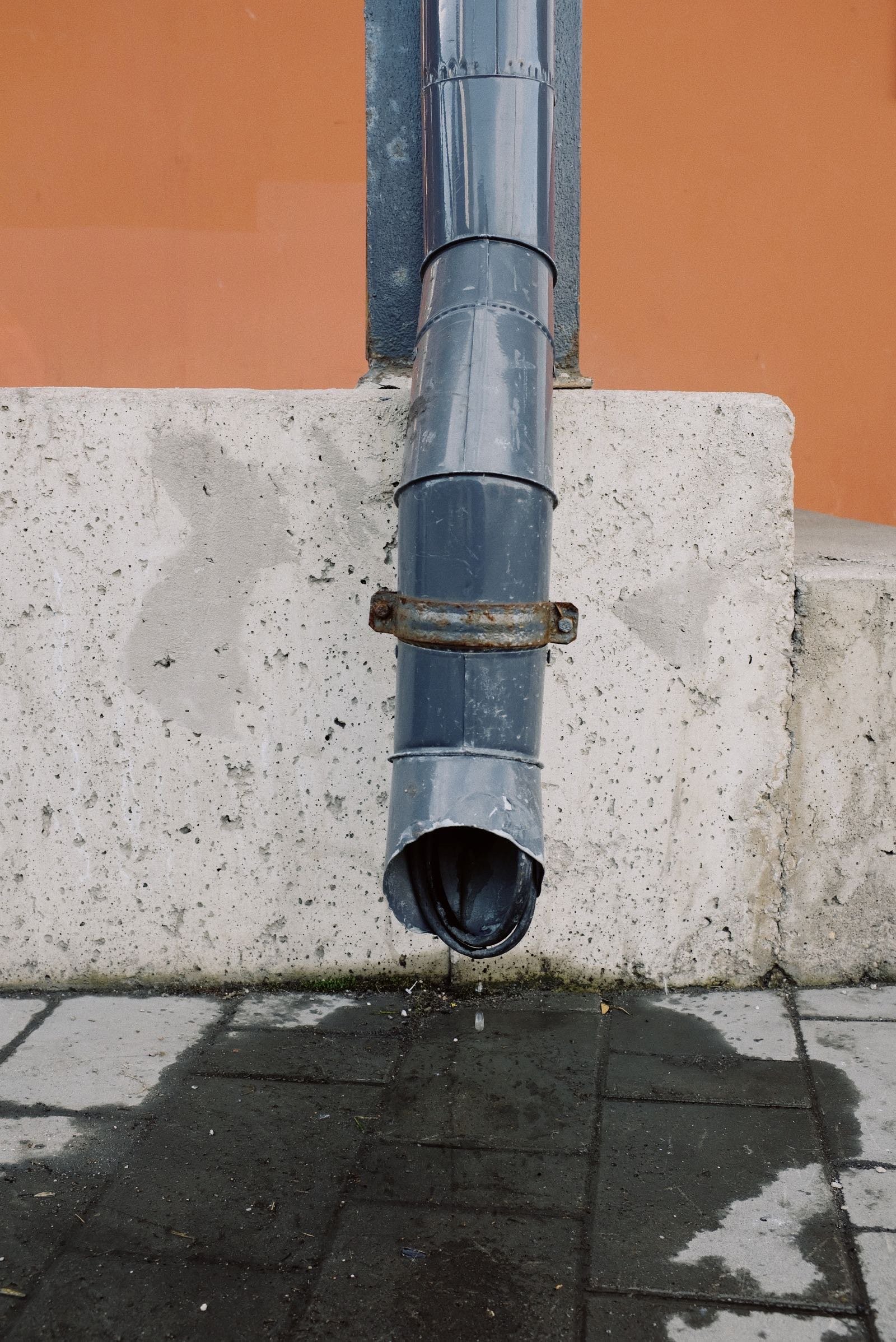 Water dripping from a downpipe onto wet paving beside a concrete wall